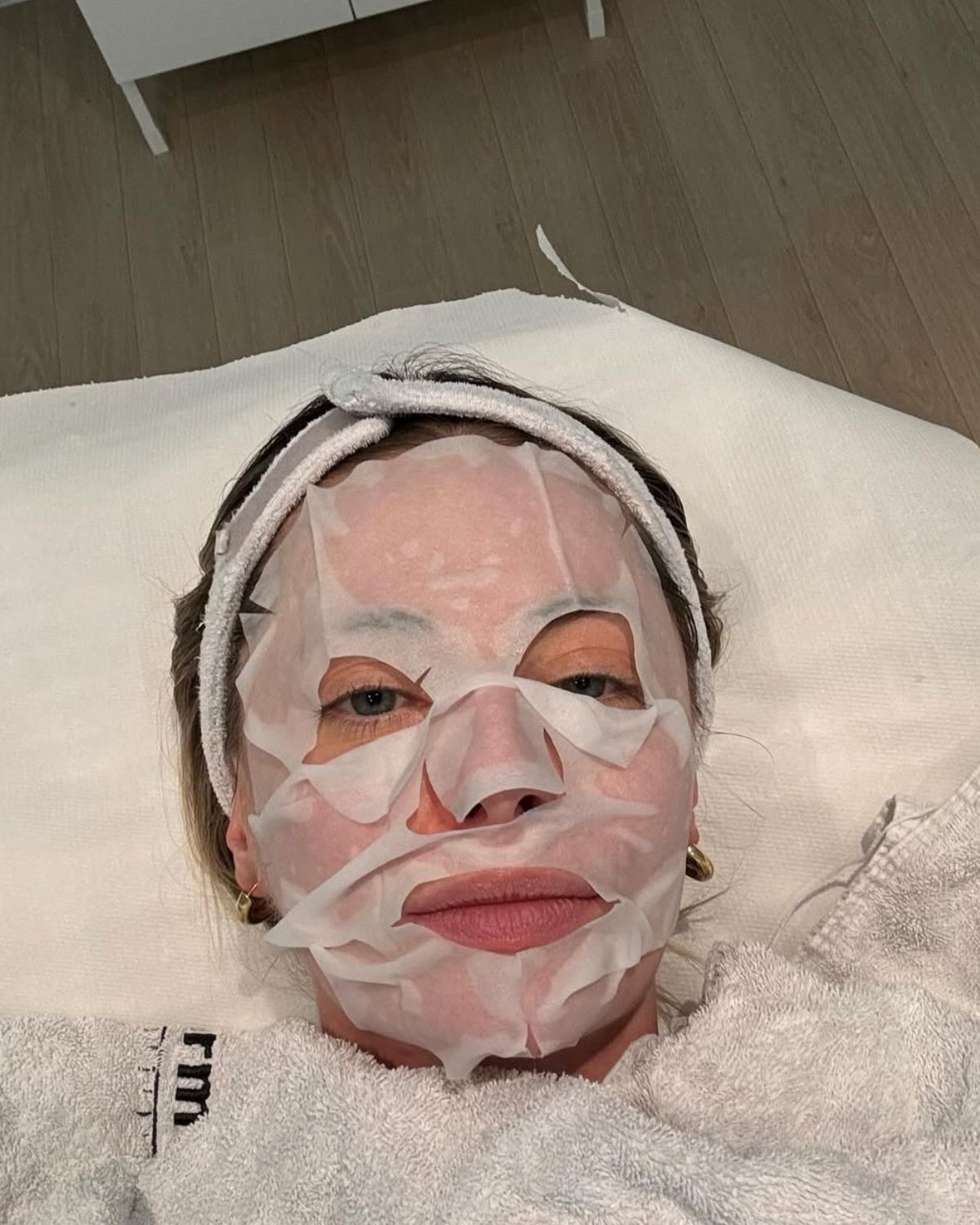 Zdjęcie zrobione jest z góry i przedstawia kobietę leżącą z nałożoną na twarz białą maską w płacie. Ma również opaskę na włosach i leży na jasnym ręczniku w relaksującej pozycji. Jest to przykład intensywnego, ale nieinwazyjnego zabiegu domowego lub w gabinecie. Ta chwila regeneracji świadczy o tym, jak priorytetowo traktuje się zabiegi pielęgnacyjne zamiast botoksu w codziennej rutynie.