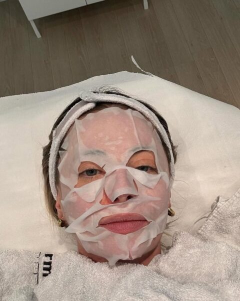 Zdjęcie przedstawia zbliżenie na kobietę leżącą na leżance zabiegowej, z maską w płachcie na twarzy i opaską na włosach, co sugeruje profesjonalny zabieg pielęgnacyjny. Jest to wizualizacja intensywnego dążenia do regeneracji i głębokiego nawilżenia cery. Kontekst zdjęcia podkreśla potrzebę eksperckiej interwencji w przypadku suchej skóry mimo nawilżania domowymi sposobami. Pielęgnacja ma na celu odbudowę i ukojenie naskórka, by przywrócić mu zdrowy blask i trwały komfort.