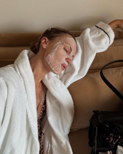 Kobieta leżąca na beżowej sofie w białym szlafroku i masce w płachcie na twarzy, relaksująca się. To idealny moment na głębokie odprężenie w ramach jej domowych rytuałów wellness, które działają lepiej niż spa w hotelu.