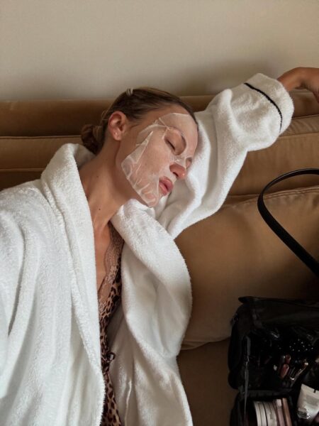 djęcie przedstawia kobietę ubraną w biały szlafrok, leżącą na beżowej sofie z częściowo nałożoną na twarz maską w płacie i zamkniętymi oczami. Widać, że jest w trakcie relaksującej chwili regeneracji, mającej na celu poprawę kondycji skóry.