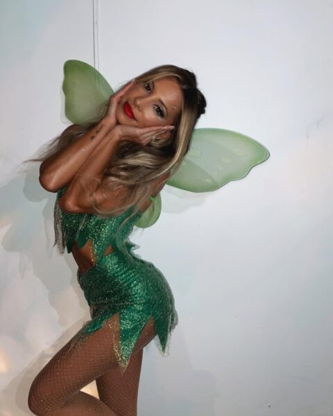 Te wróżki zmieniły naszą popkulturę. Sprawdź, którą z nich jesteś na podstawie swojego znaku zodiaku. Kobieta przebrana za Wróżkę Dzwoneczek. Ma na sobie błyszczący, zielony, dwuczęściowy kostium z postrzępionymi brzegami oraz kabaretkowe rajstopy. Za plecami ma duże, jasnozielone, półprzezroczyste skrzydła. Ma długie blond włosy, wyraźny makijaż z czerwoną szminką i pozuje, opierając dłonie na policzkach, patrząc na wprost. Tło stanowi prosta, jasna ściana.