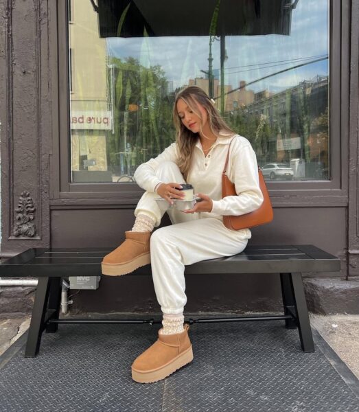 Zamienniki UGG. Kobieta siedzi na czarnej ławce przed witryną sklepową, ubrana w biały, dresowy komplet i beżowe botki na platformie. W ręku trzyma kubek, a obok niej leży brązowa torebka. Buty na platformie są stylowym przykładem zamienników UGG w wersji mini.