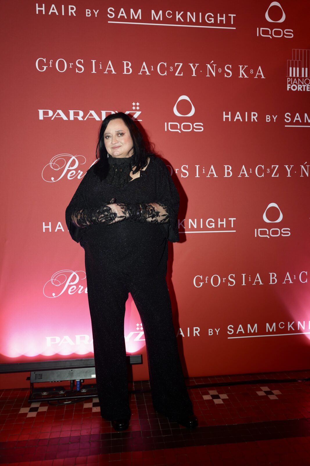 Gosia Baczyńska 2026: Osoba ubrana w czarny, połyskujący komplet z koronkowymi rękawami pozuje na tle czerwonej ścianki z logotypami marek i partnerów wydarzenia. Stoi frontalnie, z dłońmi splecionymi na wysokości brzucha, w oświetleniu podkreślającym intensywny kolor tła.