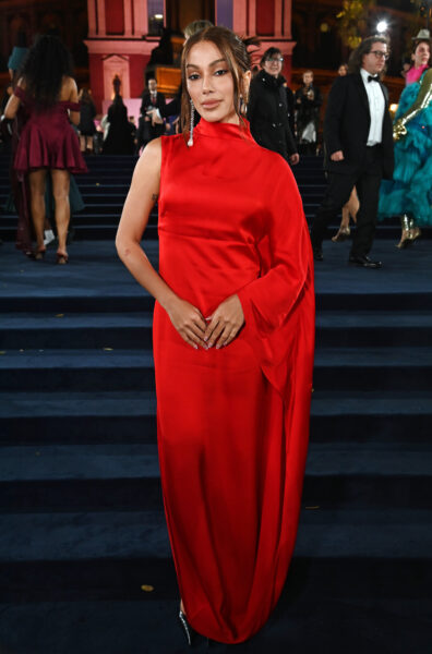 Kobieta w długiej czerwonej sukni z asymetrycznym dekoltem i drapowaną linią ramienia. Klasyczna i minimalistyczna kreacja wieczorowa w intensywnym kolorze, na tle schodów prowadzących do wejścia budynku.