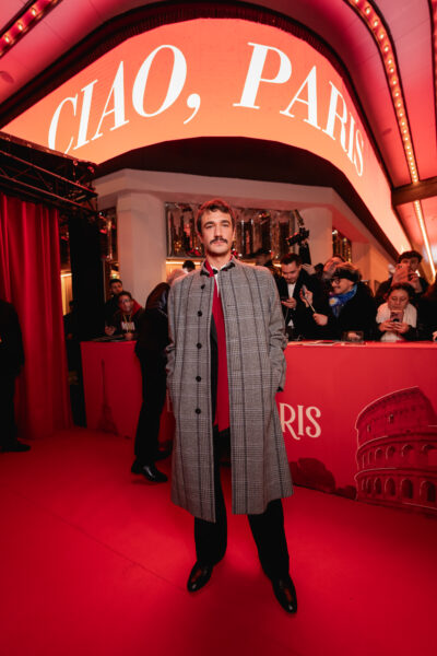 Mężczyzna stojący na czerwonym dywanie przed kinem w Paryżu, ubrany w długi szary płaszcz w kratę, czarne spodnie i bordowy krawat; w tle neonowy napis „Ciao, Paris” oraz zgromadzeni goście i fotoreporterzy podczas premiery serialu.