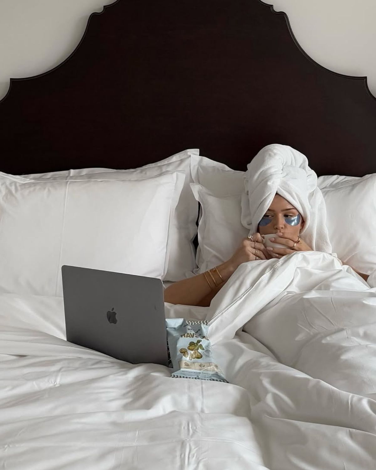 Kobieta relaksująca się w białej pościeli z ręcznikiem na głowie i płatkami pod oczami, pijąca kawę przed laptopem, planując domowe filmy na sylwestra 2025.