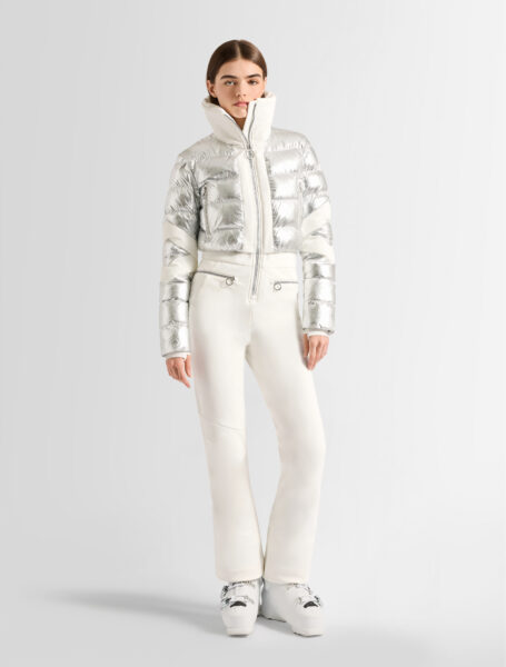 Pełna sylwetka modelki w krótkiej, srebrnej kurtce puchowej zapinanej pod samą szyję oraz dopasowanych, jasnych spodniach narciarskich; minimalistyczna stylizacja w chłodnej palecie kolorów prezentowana na czystym, studyjnym tle.