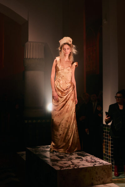 Modelka stojąca na podwyższeniu pośrodku sali w długiej, złotej sukni o drapowanym dole i odsłoniętych ramionach. Na głowie ma efektowną kokową ozdobę, a wokół panuje przyciemnione, teatralne światło z punktowymi reflektorami skierowanymi na sylwetkę.