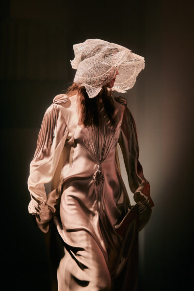 Modelka w długiej, jedwabistej sukni o strukturalnych marszczeniach i ozdobnych przeszyciach, z delikatną, koronkową woalką w formie kapelusza zasłaniającą twarz. Stoi w miękkim, scenograficznym świetle, które wydobywa połysk materiału.