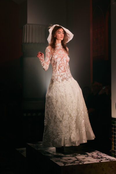 Modelka w długiej, czarnej, półprzezroczystej sukni o miękkim, lejącym kroju, rozpościerająca szerokie, zwiewne panele materiału. Stoi tyłem na podwyższeniu, z rozpiętymi ramionami, w teatralnym, kontrastowym świetle.