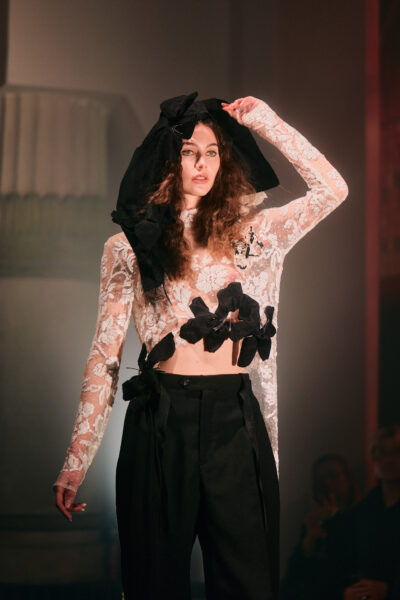 Modelka w długiej, turkusowej sukni o metalicznym połysku, z obszernym kapturem opadającym na ramiona. Stoi w świetle reflektora, które podkreśla iskrzący materiał i wyrazistą linię sylwetki.