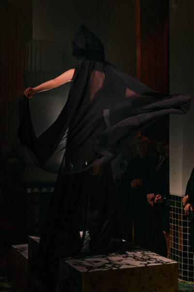 Modelka prezentuje długą, białą suknię o sylwetce koktajlowo-ślubnej, wykonaną z przezroczystej koronki z gęstym, florystycznym haftem i delikatną fakturą 3D. Góra o dopasowanym kroju z długimi rękawami i wysoką stójką subtelnie odsłania sylwetkę, przechodząc w rozkloszowany dół z warstwowego tiulu. Stylizację uzupełnia półprzezroczysta, miękka osłona głowy z tkaniny, nadająca całości teatralnego, couture’owego charakteru. Pokaz odbywa się na marmurowym podeście w nastrojowej, przygaszonej scenografii, z widownią widoczną w tle.