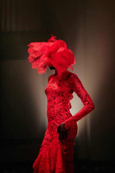 Modelka ubrana w intensywnie czerwoną, dopasowaną suknię z koronki o bogatym, kwiatowym splocie prezentuje sylwetkę inspirowaną wieczorowym haute couture. Długa forma z długimi rękawami podkreśla linię ciała, a dramatyzmu dodaje rzeźbiarska, koronkowa konstrukcja nakrycia głowy, częściowo przesłaniająca twarz. Stylizacja utrzymana w monochromatycznej czerwieni kontrastuje z mrocznym, teatralnym oświetleniem wnętrza, podkreślając ekspresyjny, performatywny charakter pokazu mody.