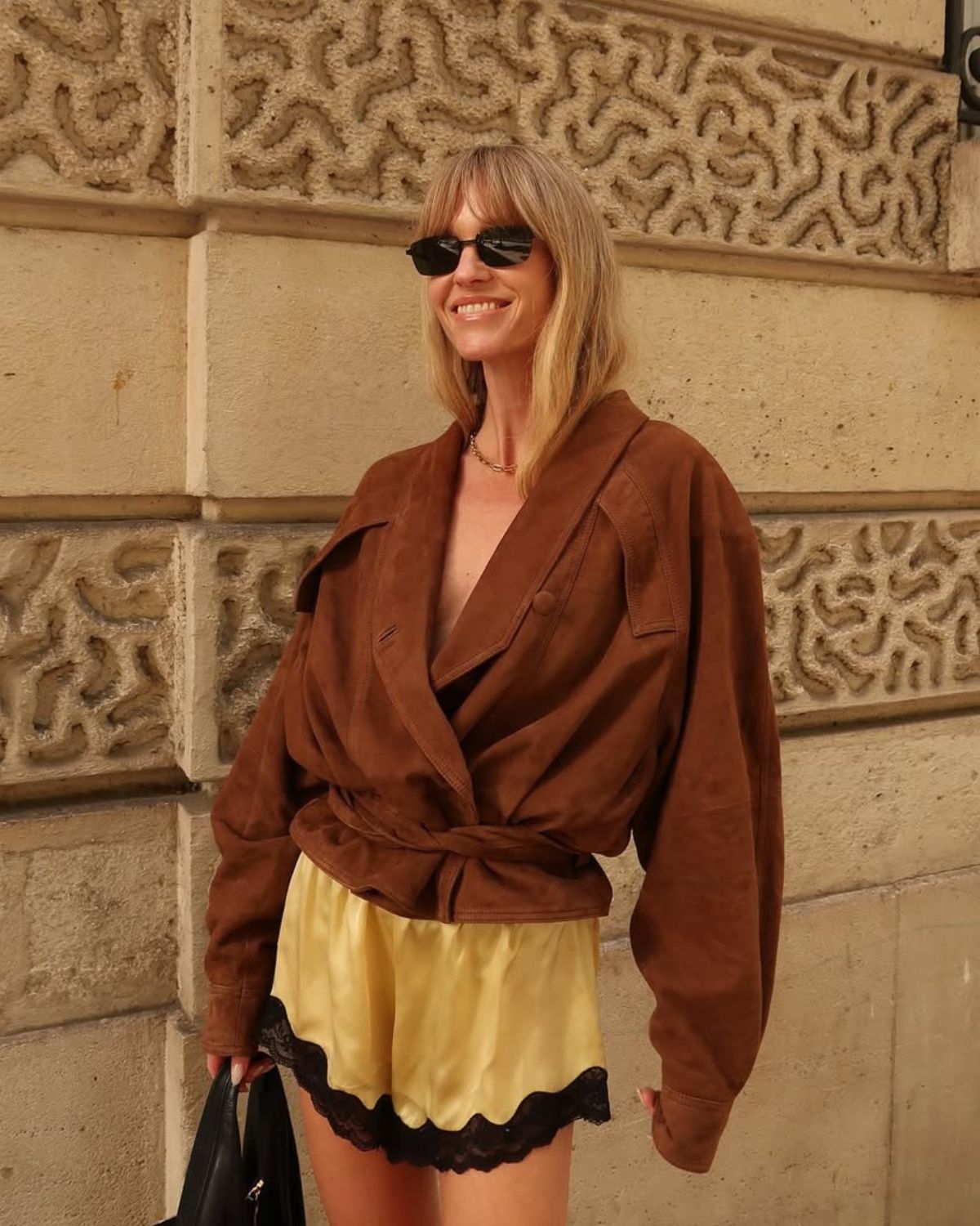 Uśmiechnięta blondynka w okularach przeciwsłonecznych, prezentująca letnią stylizację w duchu dyskretnego luksusu na tle zabytkowej elewacji. Ma na sobie brązową, zamszową kurtkę o kroju kopertowym oraz jedwabne, żółte szorty wykończone czarną koronką, co pokazuje, jak dobierać kolory ubrań do typu urody w sposób odważny i pełen klasy.