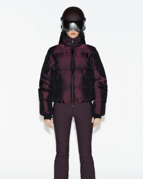 Zdjęcie przedstawia modelkę stojącą na białym tle, ubraną w komplet narciarski w kolorze głębokiego burgunda/śliwki. Ma na sobie puchową kurtkę o błyszczącym, metalicznym wykończeniu i dopasowane, obcisłe spodnie narciarskie. Twarz modelki jest całkowicie zasłonięta przez duże, futurystyczne gogle narciarskie z ciemną, lustrzaną szybą. Stylizacja jest nowoczesna i monochromatyczna.