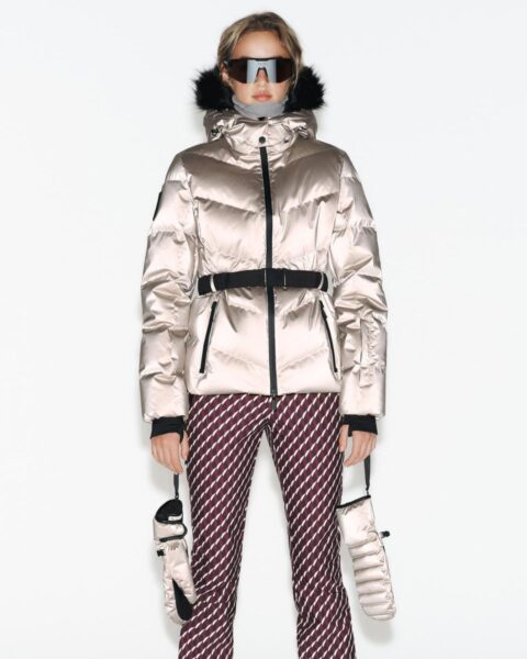 Zdjęcie przedstawia modelkę stojącą na białym tle, ubraną w modny, narciarski zestaw. Na sobie ma metaliczną, błyszczącą kurtkę puchową w kolorze jasnego szampana/beżu, przewiązaną czarnym, szerokim pasem z klamrą, oraz dopasowane spodnie narciarskie z geometrycznym wzorem w kolorze bordo i bieli. Modelka trzyma w rękach parę pasujących, puchowych rękawic. Jej twarz jest częściowo zasłonięta przez duże, czarno-lustrzane gogle narciarskie, a kurtka ma kaptur obszyty futerkiem.