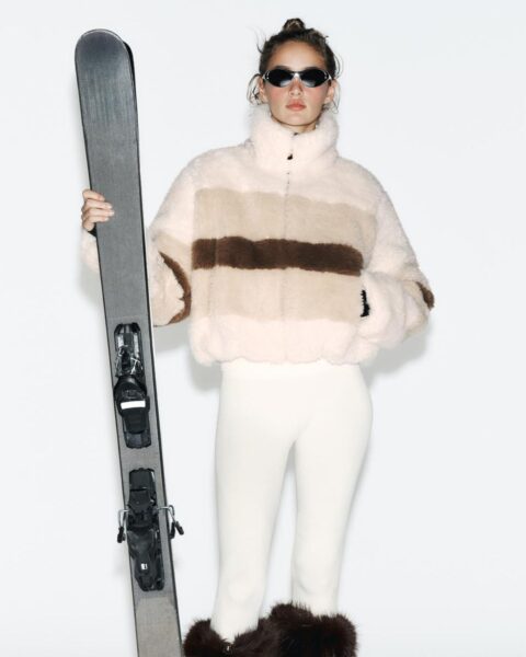 Kobieta w modnej odzieży narciarskiej z kolekcji narciarskiej Zara 2025: krótkiej kurtce typu baranek (futerko) w poziome pasy (kremowy, beżowy, brązowy) i dopasowanych, kremowych spodniach. Modelka trzyma parę nart z wiązaniami i ma na sobie wąskie okulary przeciwsłoneczne.