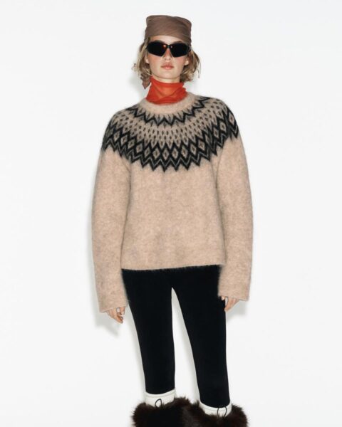 Kobieta w modnej stylizacji inspirowanej stylem apres-ski. Ma na sobie beżowy, wełniany sweter z norweskim wzorem (Fair Isle) w kolorze czarnym. Stylizację dopełniają czarne, dopasowane spodnie, futrzane, brązowe kozaki, pomarańczowa apaszka pod szyją oraz czarne okulary przeciwsłoneczne i brązowa opaska na włosach.