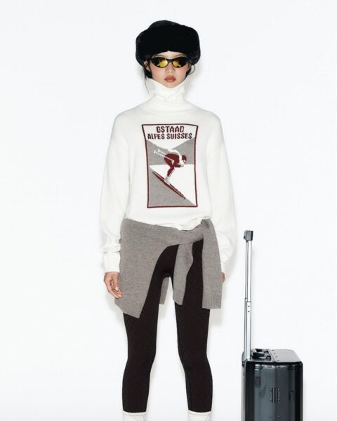 Stylizacja podróżna w klimacie retro apres-ski: Kobieta w białym, wełnianym swetrze z golfem i grafiką narciarską „Gstaad Alpes Suisses”. Ma na sobie czarne legginsy z deseniem, szary sweter przewiązany w pasie, czarny beret i futrzane śniegowce. Obok niej stoi mała, czarna walizka kabinowa.