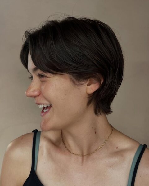 Uśmiechnięta kobieta z profilu, prezentująca krótką, ciemnobrązową fryzurę typu pixie cut w stylu lat 90. z dłuższą grzywką i odsłoniętym uchem.