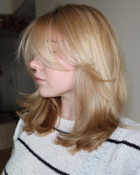 Kobieta o jasnych blond włosach w fryzurze typu warstwowy bob (butterfly cut) z dłuższą grzywką na boki. Włosy sięgają do ramion, są lekko wywinięte na końcach i mają miodowy odcień. Modelka jest zwrócona profilem, ubrana w biały sweter w czarne paski.