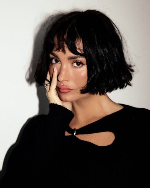 Portret kobiety o ciemnych, krótkich włosach typu bob z grzywką. Modelka ma na sobie czarną bluzkę z ozdobnym wycięciem przy dekolcie, opiera dłoń o policzek i patrzy bezpośrednio w obiektyw. Zdjęcie charakteryzuje się mocnym kontrastem i wyraźnym cieniem na jasnym tle, podkreślającym rysy twarzy i elegancki makijaż z matową, różową szminką.