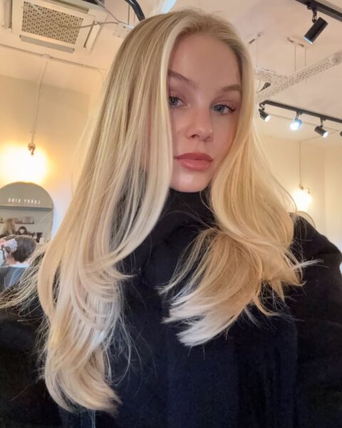 Kobieta o bardzo długich, gęstych, blond włosach w odcieniu butter blonde (maślany blond). Fryzura jest idealnie wystylizowana z widocznym, subtelnym cieniowaniem warstwowym (tzw. layering), dodającym objętości. Kobieta ma na sobie czarny, miękki golf lub sweter. Ujęcie wykonane w nowoczesnym salonie fryzjerskim, z widocznym lustrem i oświetleniem punktowym.