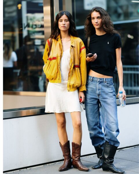 Dwie młode kobiety na ulicy w Nowym Jorku. Po lewej: Biała sukienka mini, musztardowo-żółta kurtka bomberka ze sportowymi detalami i kowbojki w kolorze gorzkiej czekolady. Po prawej: Czarny, krótki T-shirt polo z logo i luźne jeansy z niskim stanem, wpuszczone w czarne, wysokie oficerki. Inspiracja stylem street fashion Nowy Jork.