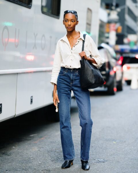 Kobieta w stylizacji street fashion Nowy Jork. Klasyczne, granatowe jeansy z wysokim stanem połączone z beżową, luźną koszulą z podwiniętymi rękawami. Look uzupełniają czarne botki na niskim obcasie, czarne, wąskie okulary przeciwsłoneczne i duża, czarna, pleciona torebka typu tote. Elegancki minimalizm w modzie ulicznej.