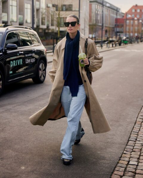 Elegancka stylizacja street fashion w Kopenhadze. Kobieta ma na sobie klasyczny, beżowy trencz, granatowy sweter i długi, ciemny szal. Dół stanowią szerokie, jasnoniebieskie jeansy, a całość uzupełniają czarne kanciaste okulary przeciwsłoneczne. W dłoni trzyma zielony koktajl. Moda skandynawska jesień – inspiracja na stylowy płaszcz.