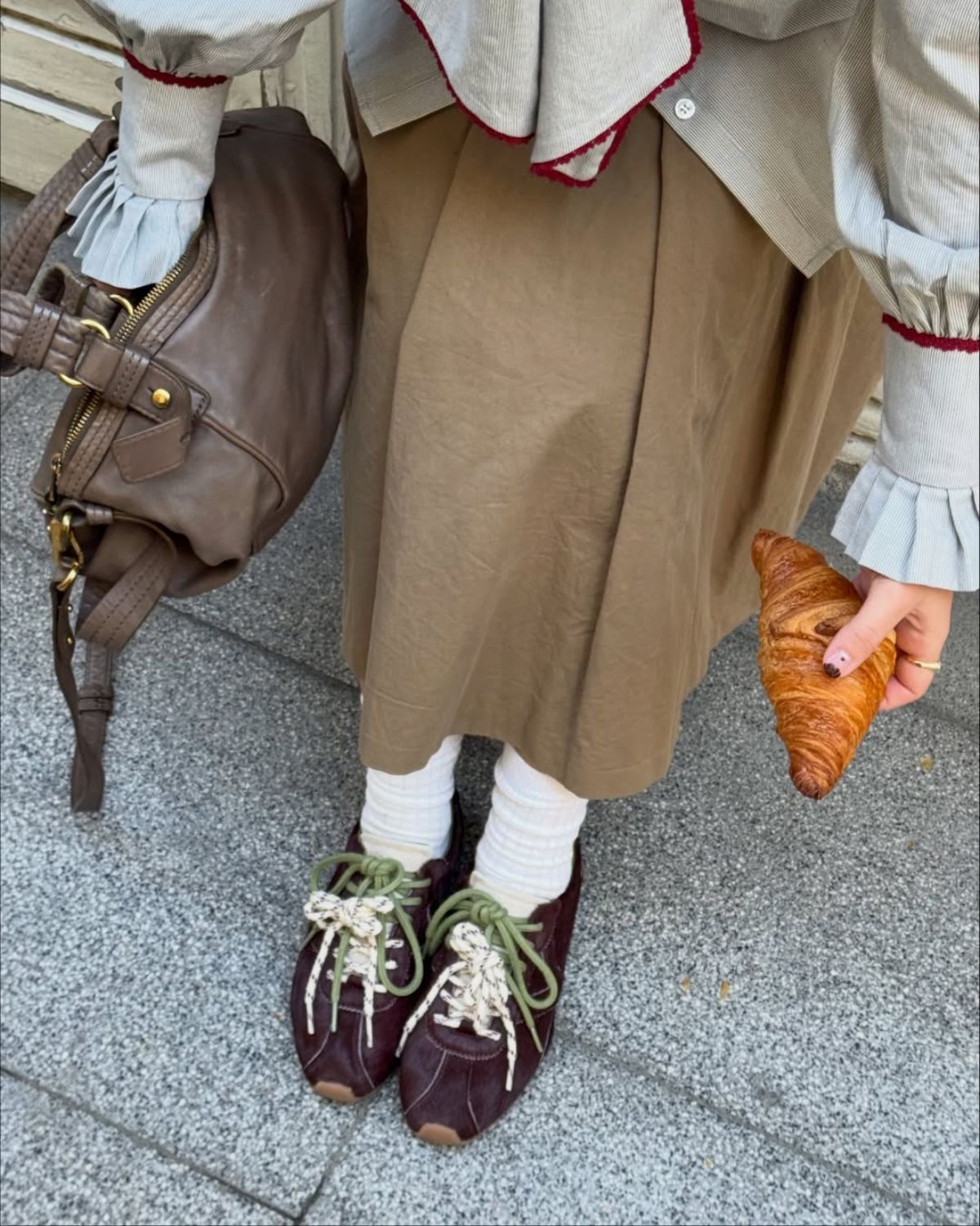 Modne sneakersy 2025 w kolorze bordowym ze sznurówkami w odcieniach zieleni i ecru, zestawione z białymi skarpetkami, beżową spódnicą midi i koszulową bluzką z bufiastymi rękawami. W tle widać chodnik, na którym stoi modelka. Modelka trzyma w dłoni rogalika, a na ramieniu ma brązową skórzaną torebkę. Stylizacja łączy estetykę cottagecore z miejskim szykiem.
