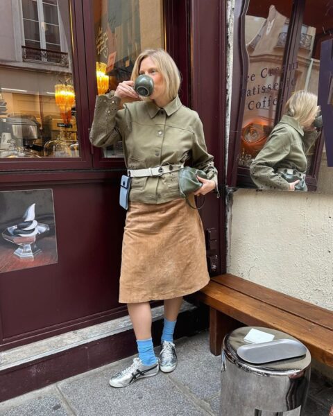 Kobieta o blond włosach, w stylu streetwear, pijąca kawę na zewnątrz kawiarni. Ma na sobie oliwkową, krótką kurtkę z haftowanymi rękawami, brązową zamszową spódnicę do kolan, biały pasek i metaliczne srebrne sneakersy, widoczne z niebieskimi skarpetkami. Trzyma małą zieloną torebkę. Stylowa inspiracja na jesienny dzień w mieście, np. w Paryżu.