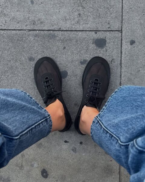 Widok na stopy w czarnych, siateczkowych (lub tekstylnych), minimalistycznych sneakersach z czarnymi sznurówkami i na czarnej podeszwie. Buty są zestawione z podwiniętymi, niebieskimi jeansami. Zdjęcie zrobione z góry, na tle szarego, betonowego chodnika.