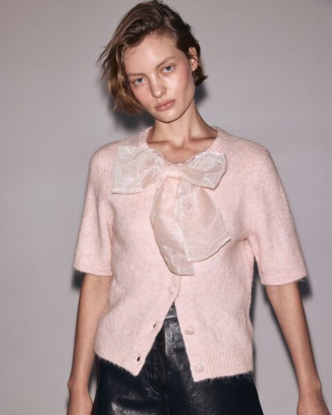 Elegancka stylizacja na Święta lub Nowy Rok: modelka w jasnoróżowym, puszystym, krótkim kardiganie z krótkimi rękawami, ozdobionym dużą, transparentną kokardą w kolorze nude przy dekolcie. Dół to czarne, skórzane szorty. Najładniejsze swetry świąteczne – inspiracja na stylowy, choć nieoczywisty, świąteczny look.