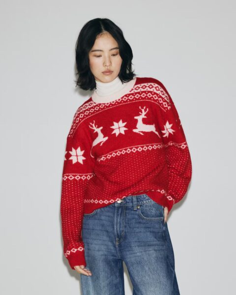 Modelka w czerwonym, wełnianym swetrze świątecznym typu Ugly Sweater, zdobionym norweskim wzorem z białymi reniferami i płatkami śniegu. Pod spodem biały golf. Dół stylizacji stanowią modne, szerokie jeansy wide leg w niebieskim odcieniu. Najładniejsze swetry świąteczne w klasycznej, lecz stylowej odsłonie.
