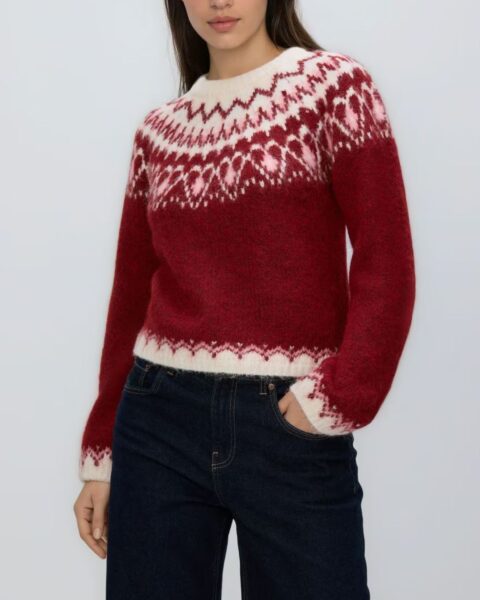 Modelka w półzbliżeniu, ubrana w klasyczny, bordowy sweter świąteczny zdobiony białymi, norweskimi wzorami: gwiazdami, płatkami śniegu i reniferami. Pod szyją nosi srebrny, warstwowy naszyjnik. Stylizacja idealna dla kobiet z klasą na zimowe okazje.