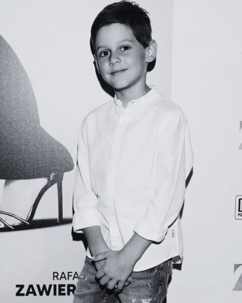 Chłopiec, Nikodem Marecki, w białej koszuli na premierze filmowej. Stoi uśmiechnięty na tle plakatu z elementem damskiego obuwia. Nikodem Marecki kim był.