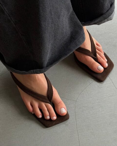 Zdjęcie prezentuje blask i perfekcyjny wygląd kremowego (mlecznego) pedicure, idealnego dla kobiet z klasą. Paznokcie u stóp są pomalowane eleganckim odcieniem kremowej bieli lub bardzo jasnego beżu. Stopy ubrane są w modne, kwadratowe klapki typu flip-flop z szerokim, brązowym paskiem. W tle widać postrzępione, ciemnoszare nogawki szerokich dżinsów i jednolitą, szarą podłogę. Jest to zwięzły i szykowny look na lato.