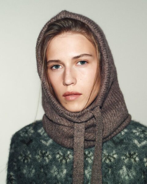 Portret blondynki w beżowo-brązowej, wełnianej kominiarce (balaklavie) z długimi wiązaniami, zestawionej z zielonym swetrem w norweski wzór. Stylowe i ciepłe nakrycie głowy idealne na **prezenty na gwiazdkę z Zary** dla kobiety ceniącej modę i funkcjonalność.