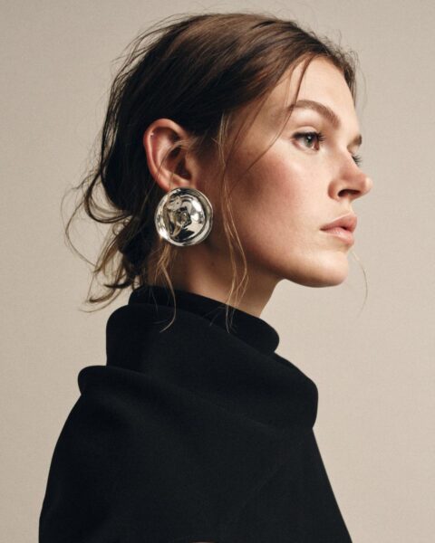 Portret modelki w czarnym golfie i dużym, okrągłym, srebrnym kolczyku. Kolczyk ma rzeźbioną, wypukłą formę, dodając stylizacji luksusowego, architektonicznego charakteru. Modne akcesoria damskie.