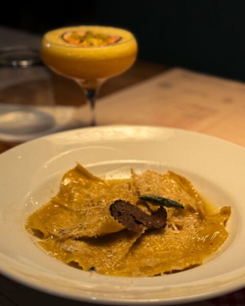 Włoskie ravioli z kozim serem i truflą, podane na eleganckim białym talerzu w nastrojowym świetle restauracji; w tle rozmyty koktajl z marakują, tworzący luksusowy klimat wieczornej kolacji.