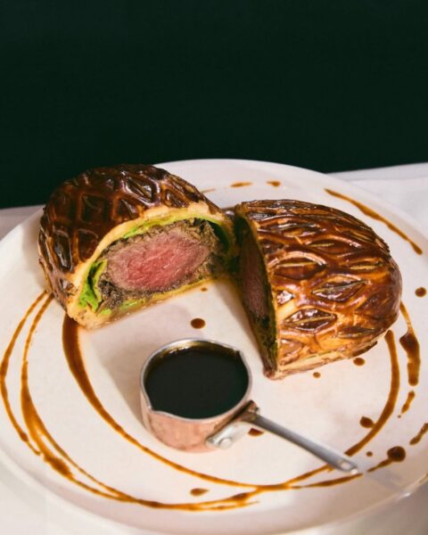 Klasyczna polędwica Wellington z różowym wnętrzem, otulona aromatycznym portobello i złocistym ciastem francuskim, podana z gęstym sosem na białym talerzu; wykwintne danie serwowane przez restauracje otwarte w Nowy Rok 2026 w Warszawie.