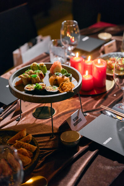 Elegancko nakryty stół podczas kolacji MIUMAG x SEN, z centralnie ustawionym talerzem pełnym sushi na metalowym stojaku, otoczonym świecami w czerwonym kolorze, kieliszkami i winietką z imieniem gościa „Oliwia Bosowska”.