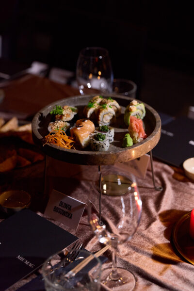 Zbliżenie na półmisek sushi stojący na podwyższeniu pośród nakryć stołowych i kieliszków; w kadrze widoczna winietka „Oliwia Bosowska” oraz przyciemnione, nastrojowe światło kolacji.