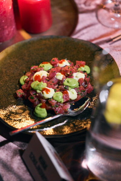 Talerz z tataki z tuńczyka podanego z zielonymi i białymi sosami w formie dekoracyjnych kropli; obok czerwone świece i kieliszki, tworzące atmosferę eleganckiej, kameralnej kolacji.