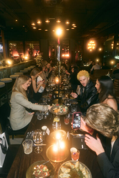 Długa, elegancka kolacja MIUMAG x SEN przy świecach, z gośćmi siedzącymi po obu stronach stołu, fotografującymi potrawy i rozmawiającymi w nastrojowej, klubowej przestrzeni.