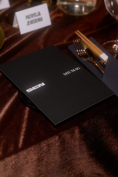 Czarna karta menu z logo SEN i MIUMAG leżąca na satynowym, brązowym obrusie, obok sztućców, pałeczek i winietki z nazwiskiem gościa.