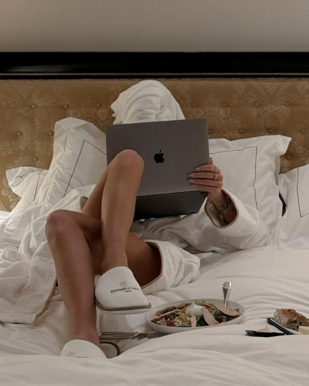 Kobieta w białym szlafroku i ręczniku na głowie relaksuje się w luksusowym łóżku hotelowym, jedząc kolację z room service i oglądając skandynawskie seriale kryminalne na laptopie.