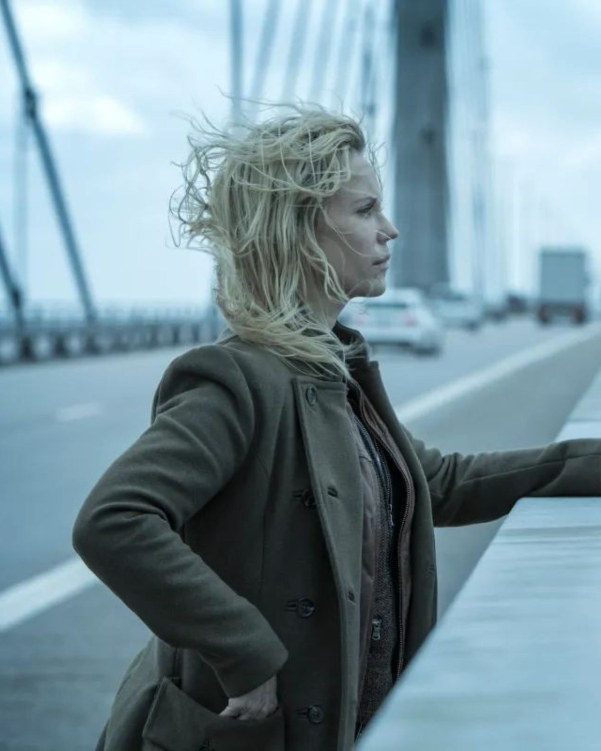 Kobieta w beżowym, wełnianym płaszczu z kołnierzem, o blond włosach rozwianych przez wiatr, stoi na moście, opierając się o barierkę. W tle widać drogę i ruch uliczny oraz szarą, mglistą aurę nad wodą. Jej mina jest poważna, a scena ma mroczny, skandynawski klimat. To zdjęcie idealnie oddaje atmosferę, jaką cechują się skandynawskie seriale na zimę.