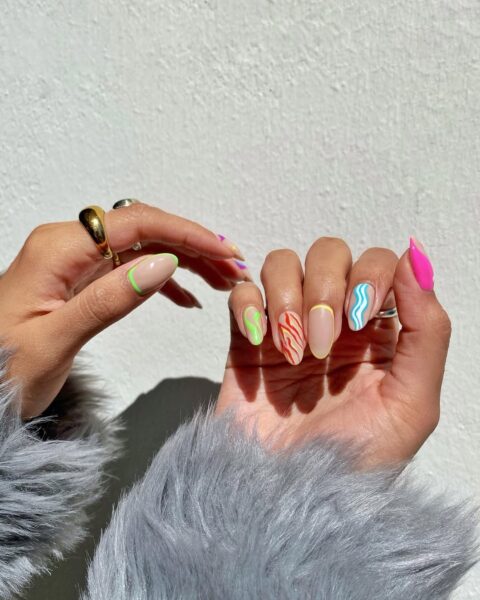 Zdjęcie pokazuje dłonie z asymetrycznym, kolorowym manicure, w którym każdy paznokieć ma inny, dynamiczny wzór w intensywnych odcieniach. Połączenie nieregularnych linii, kontrastów kolorystycznych i błyszczącej biżuterii idealnie wpisuje się w trend glitchy glam.